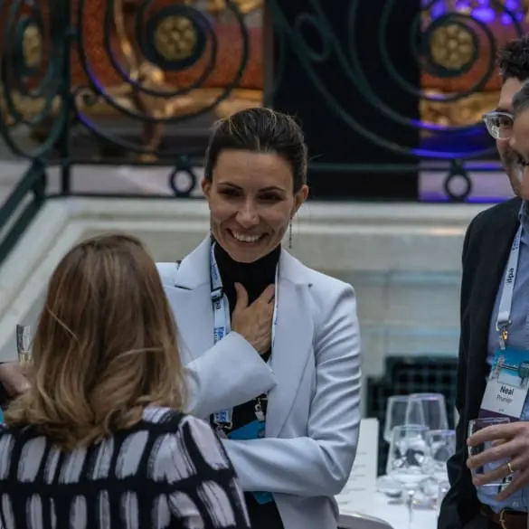 A woman wearing a white suit jacket smiles and takes to another woman in a printed shirt with her back to the camera. The smiling woman has a lanyard around her neck.